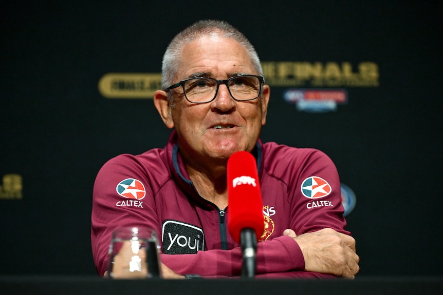 A man in a Brisbane Lions polo wearing glasses speaks into a microphone