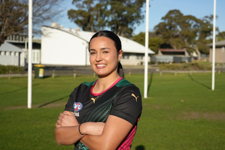 A young woman with dark hair standing with her arms crossed smiling in front of footy goals.