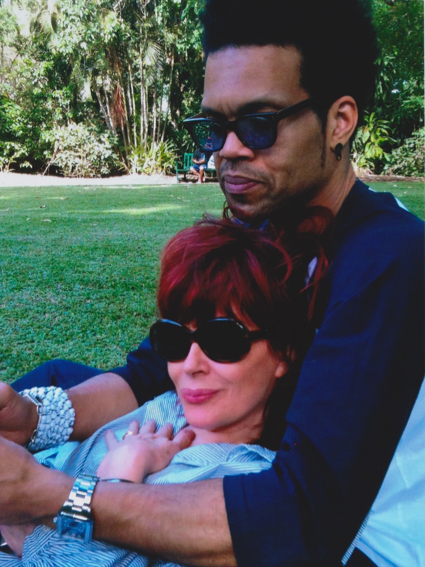 Chrissy Amphlett wears sunnies and bright blue shirt and lays in husband Charley Drayton's lap.