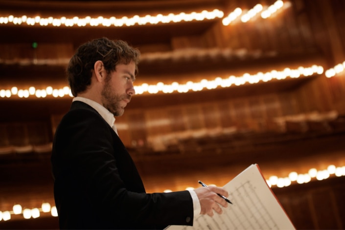 Side-on shot of Bryce Dessner in tuxedo looking over a music score sheet with blurred concert hall lights in the background.
