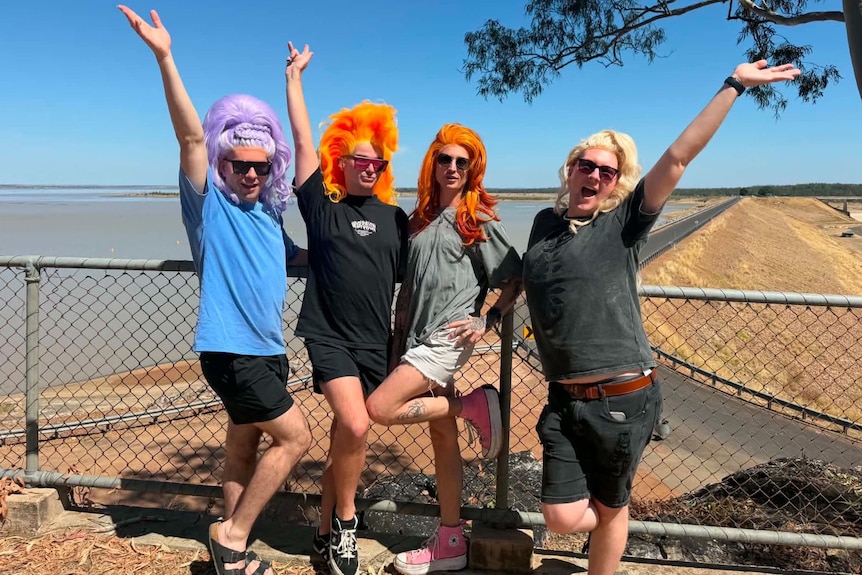 Four men in drag wigs pose in front of Emerald's Fairbairn Dam, with wide horizon and country road.