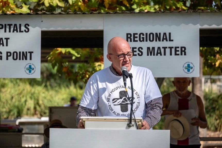 A man stands at a microphone by a pulpit next to an outdoor shelter and trees.