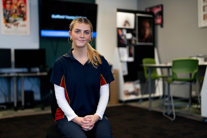 Ella sitting on a chair in a classroom with computers and TV behind her, wearing a school t-shirt and smiling.
