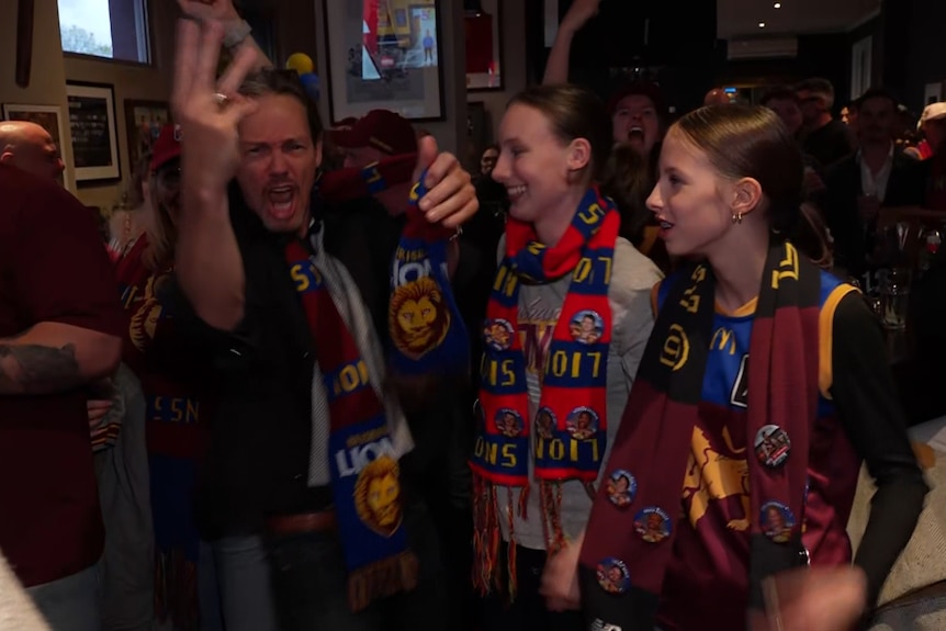 People in a dark bar, many in Brisbane Lions supporter gear, with one man holding three fingers in the air.