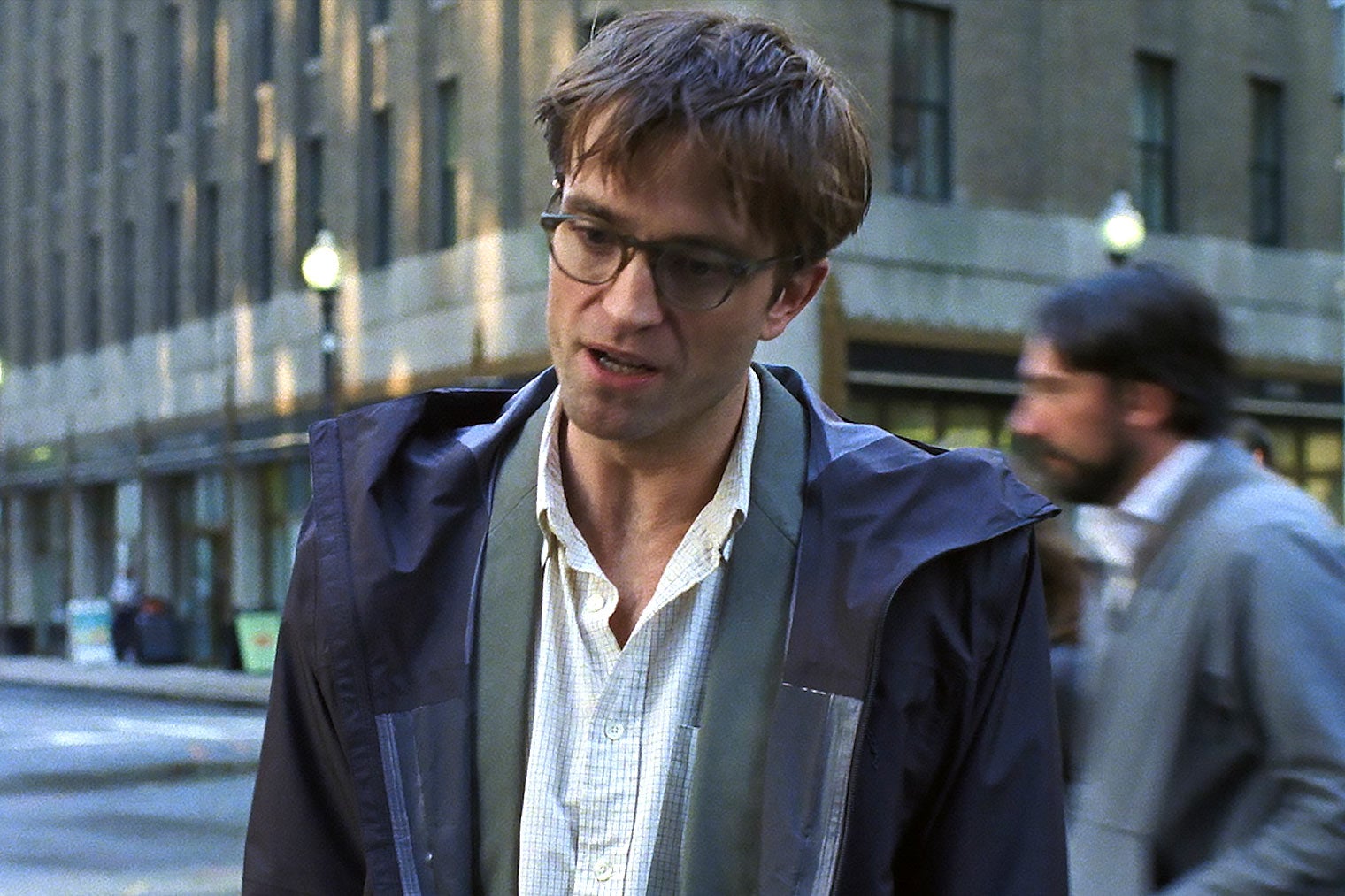 A thirtysomething man in glasses and a raincoat standing on a street and looking down.