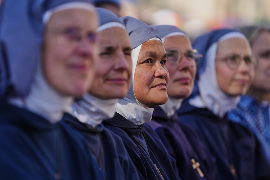 Six women wearing blue nun outfits sit side by side looking off into the distance.