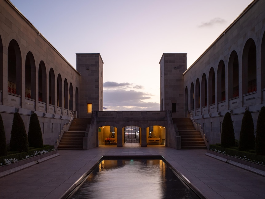 A flame sitting in a long reflecting pool. Above it is the sandstone building of the war memorial at dusk.