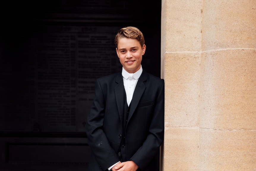 A boy stands in a suit leaning on a sandstone pillar.