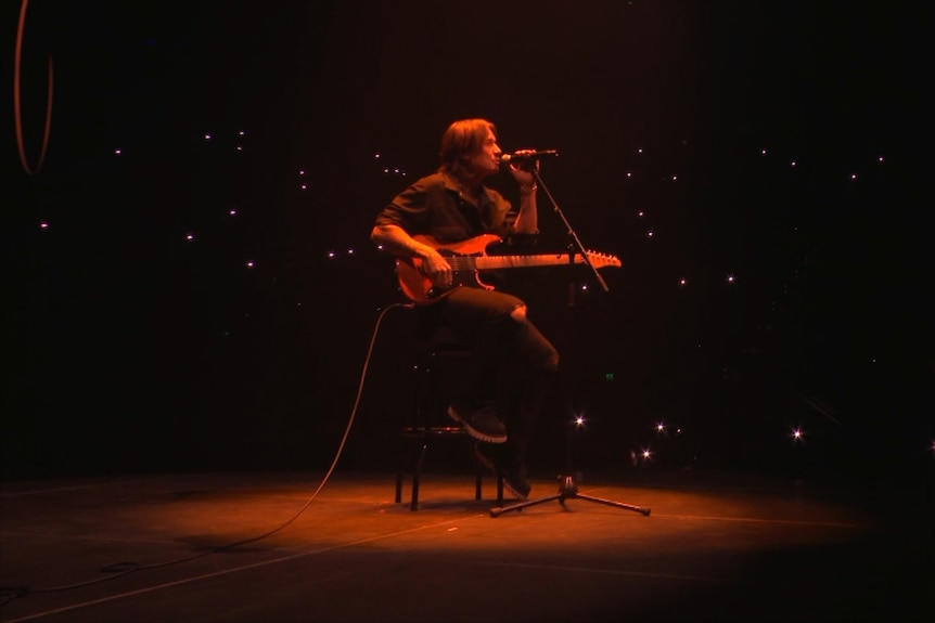 Keith Urban sits on a stool on stage and plays guitar.