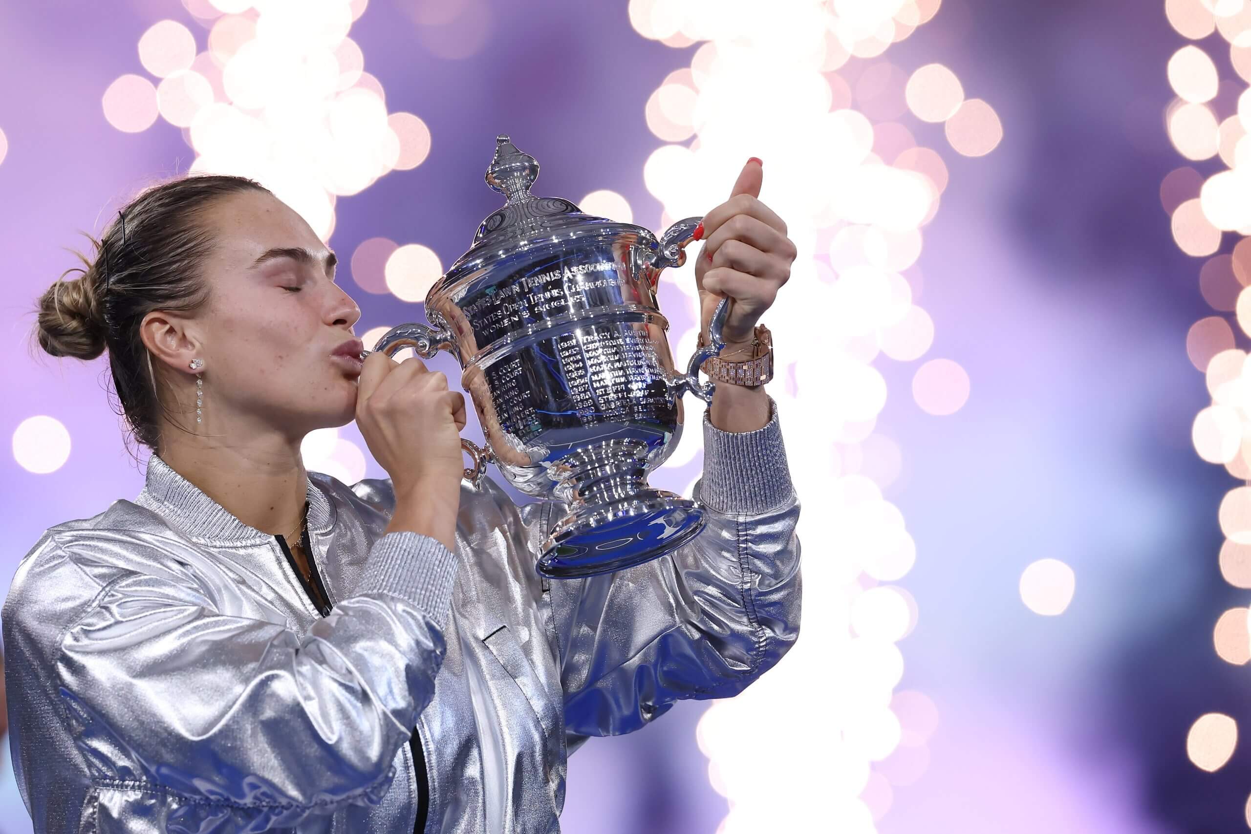 Aryna Sabalenka kisses the U.S. Open singles trophy wearing a silver jacket.