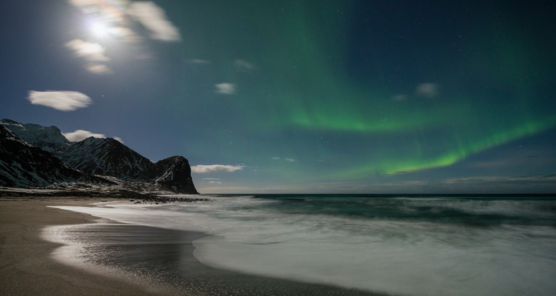 Aurora borealis lights up the night sky above a snowy mountain and beach shoreline, with waves gently washing onto the sand.