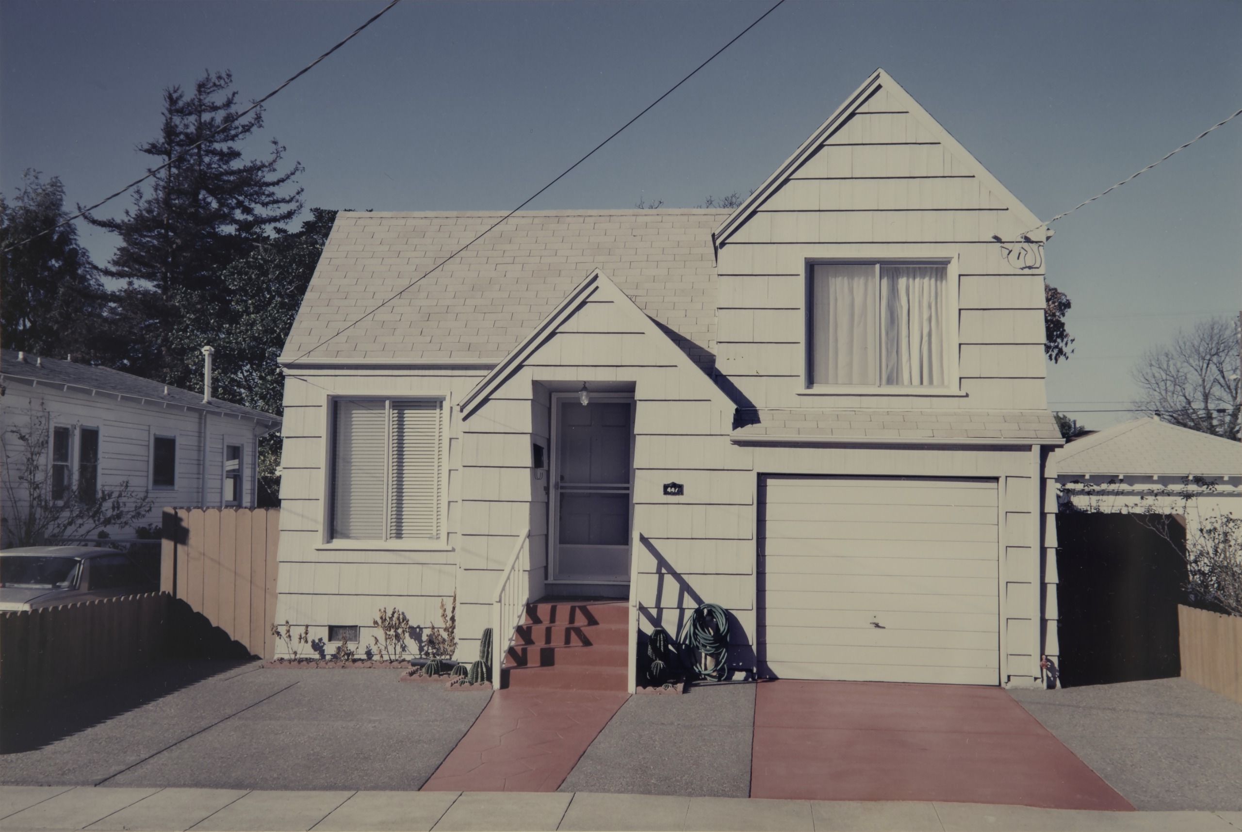 A colour photograph of a pale two-storey house with a red driveway and pathway, a garage, and a car parked beside a wooden fence.