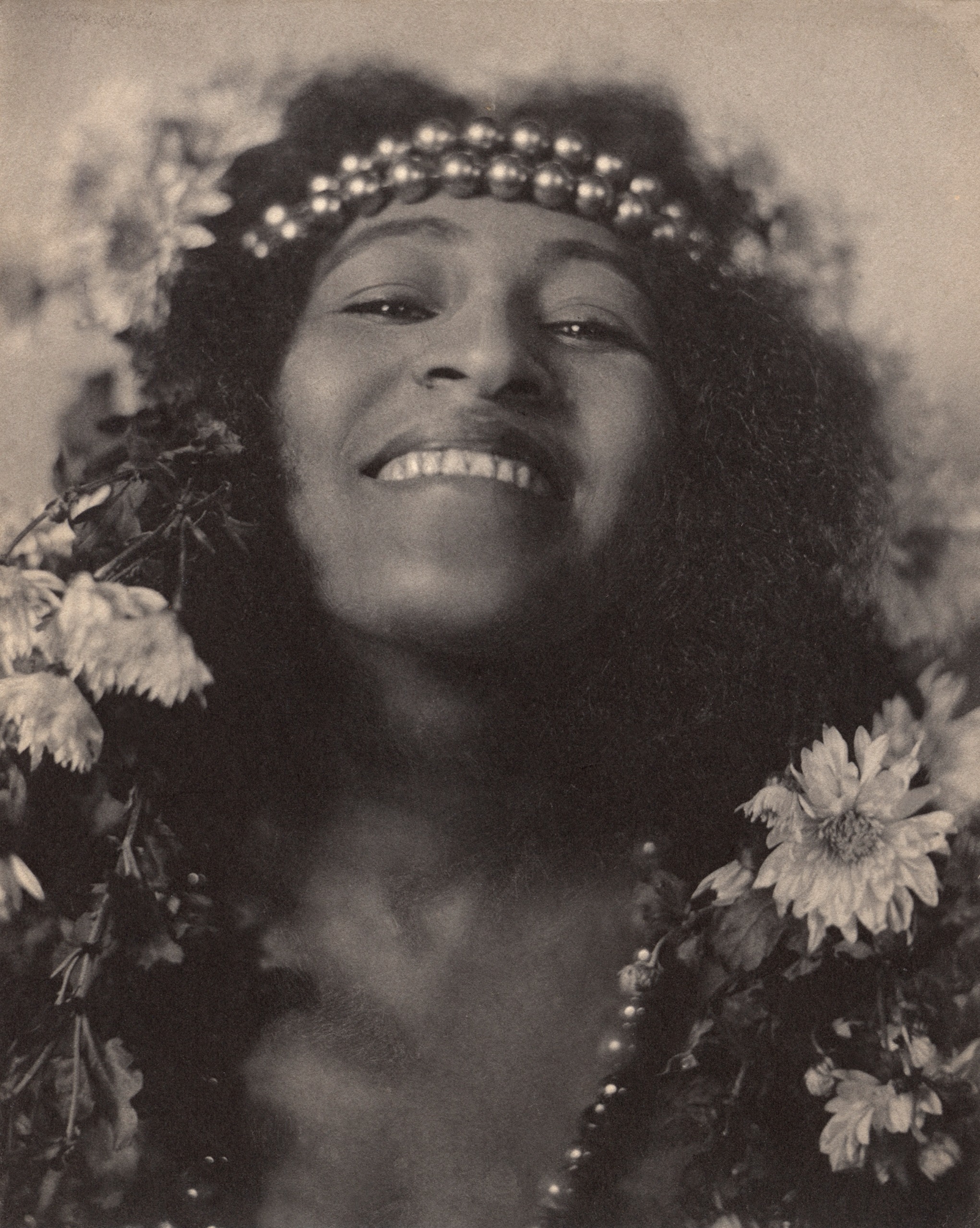 A softly focused close-up portrait of a smiling woman crowned with flowers and a beaded headband, her hair framed by blossoms.