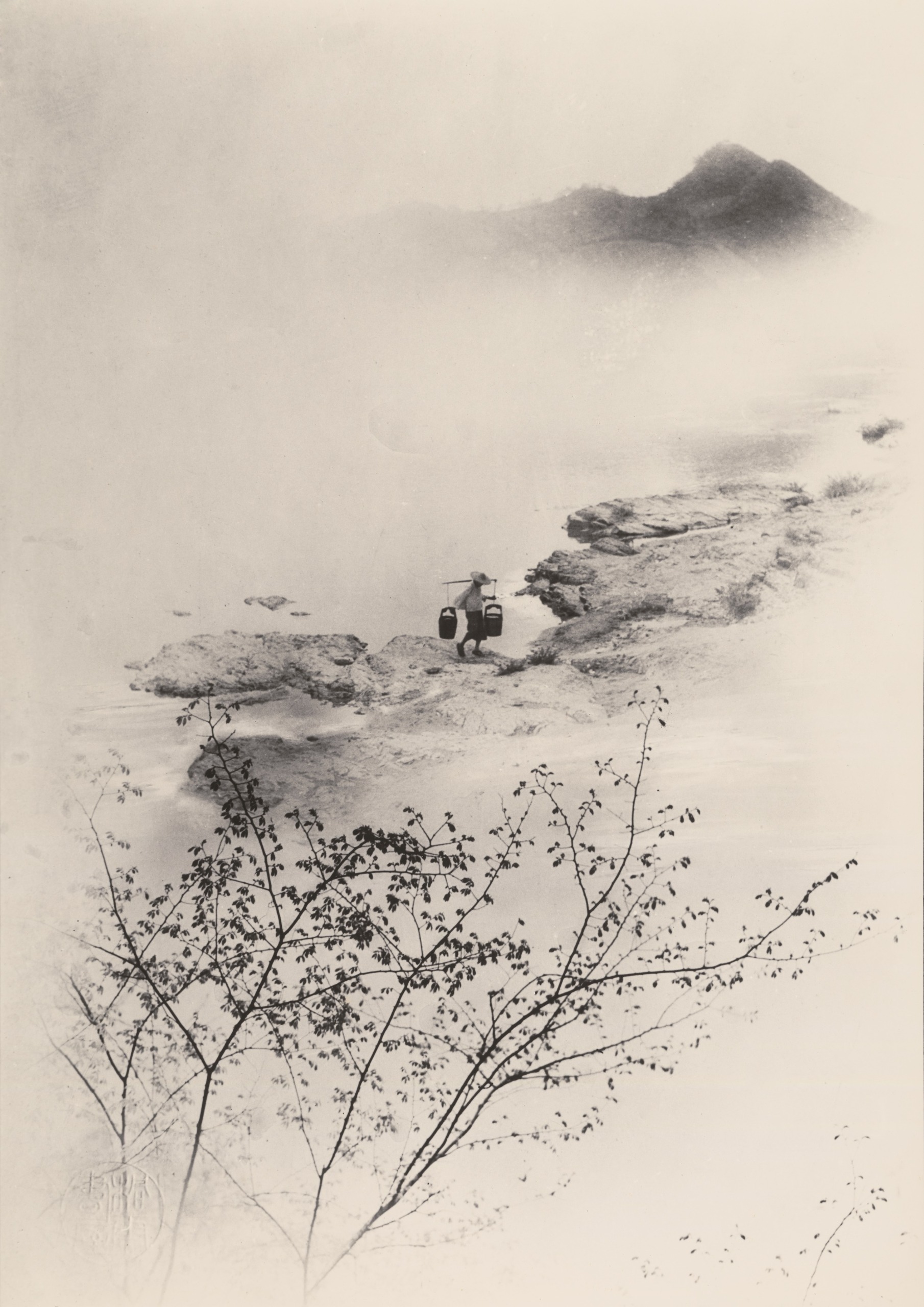 A misty riverside landscape in a soft, ink-wash style, with a lone figure in a wide-brimmed hat carrying baskets on a shoulder pole along a rocky shore beneath a distant mountain.