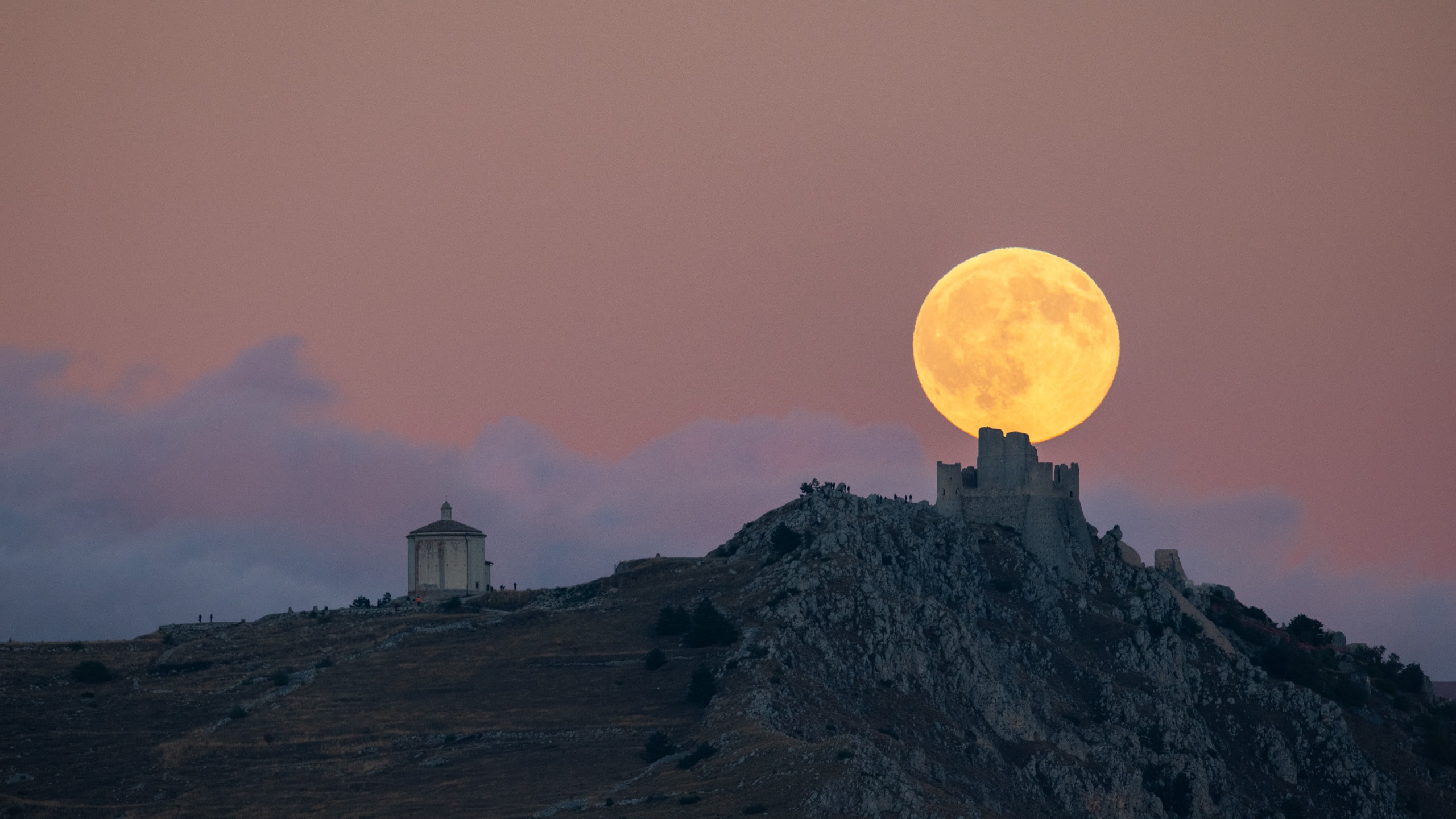 A large golden full moon rises behind the silhouette of Rocca Calascio Castle and the Church of Santa Maria della Pietà in Calascio, Italy.