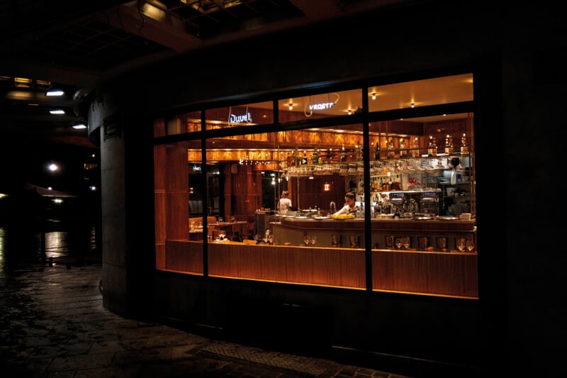 A brightly lit bar interior is viewed through a large window at night, showing a bartender working behind a wooden counter.