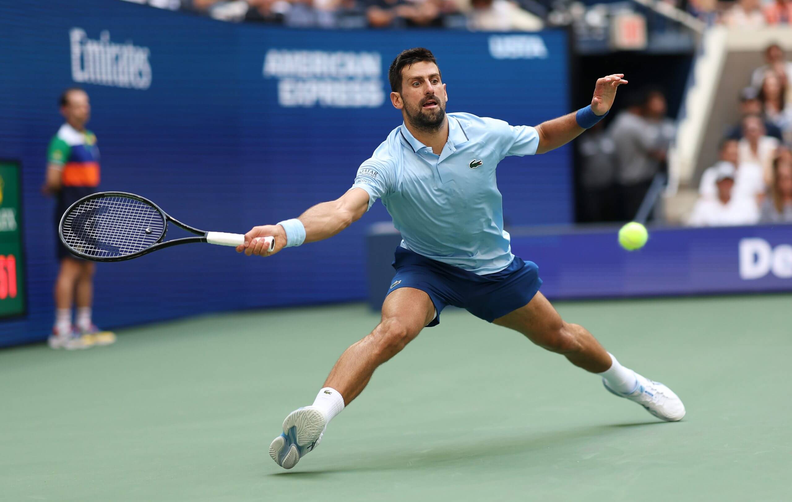 Novak Djokovic slides out to hit a forehand on the green acrylic behind a blue tennis court at the U.S. Open.