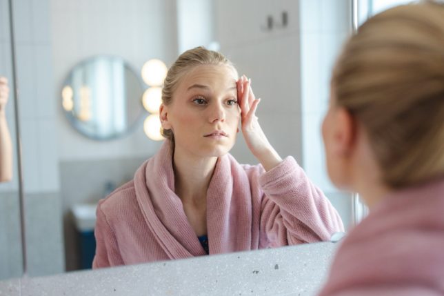 Young woman wearing a pink robe examines her face in a bathroom mirror, performing a gentle facial massage as part of her morning skincare routine and self-care ritual.