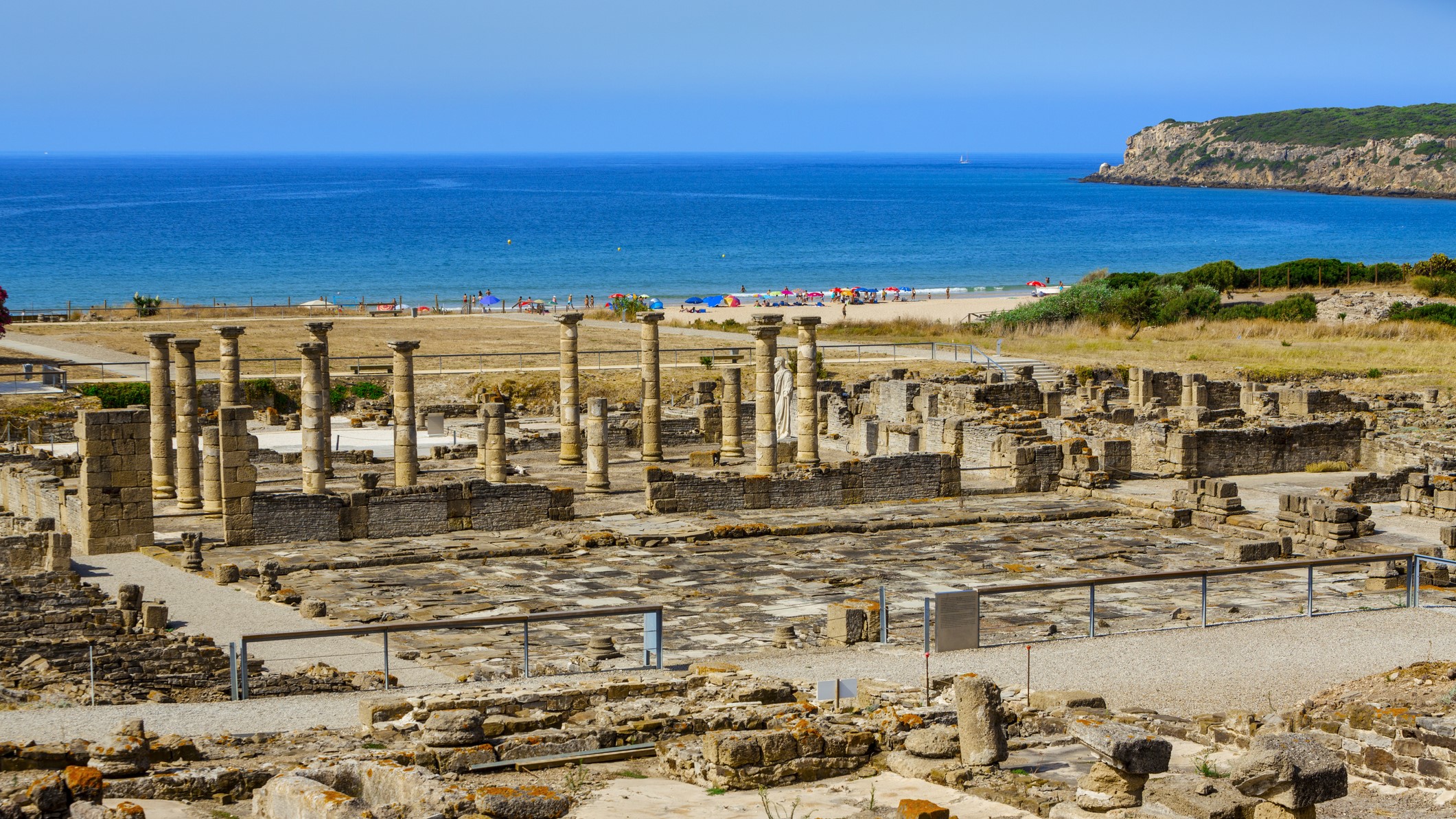 a large ruin containing numerous columns is in the foreground of the image with a beach in the background lined with parasols and people.