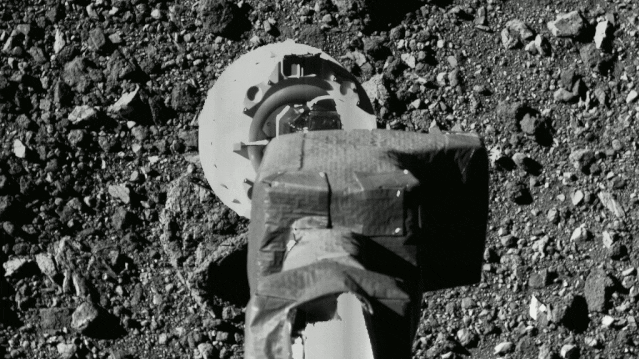 A spacecraft's arm above a moving ground. It's black and white and the ground looks pebbled and rocky.