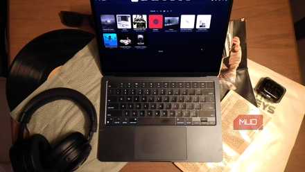 A MacBook playing music on a desk cluttered with music CDs, a vinyl record, and headphones