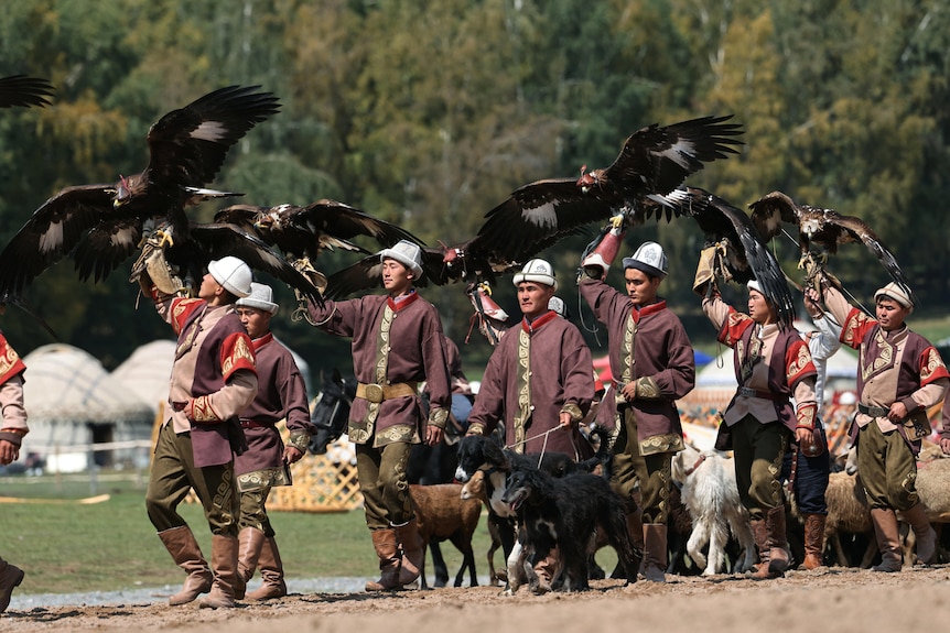 Men wearing traditional Central Asian dress, holding eagles and leading dogs.