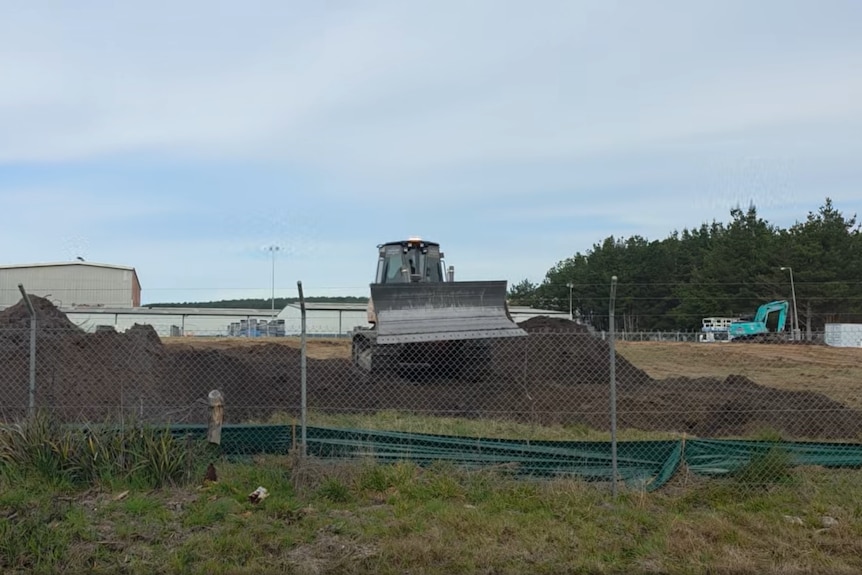 An excavator on a pile of dirt, behind a fence.