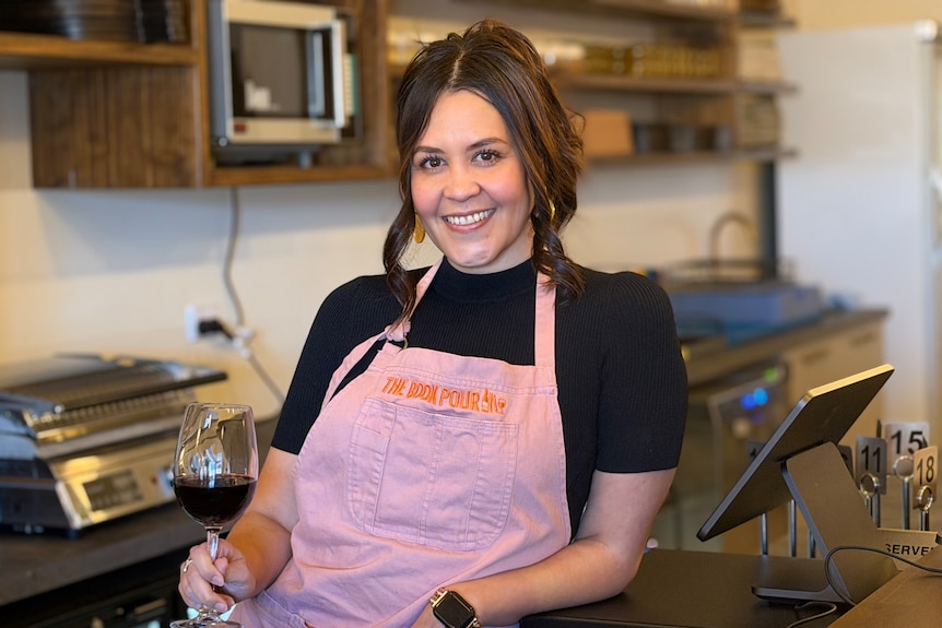 A woman leans on a bar near a cash register with a glass of wine in her hand.