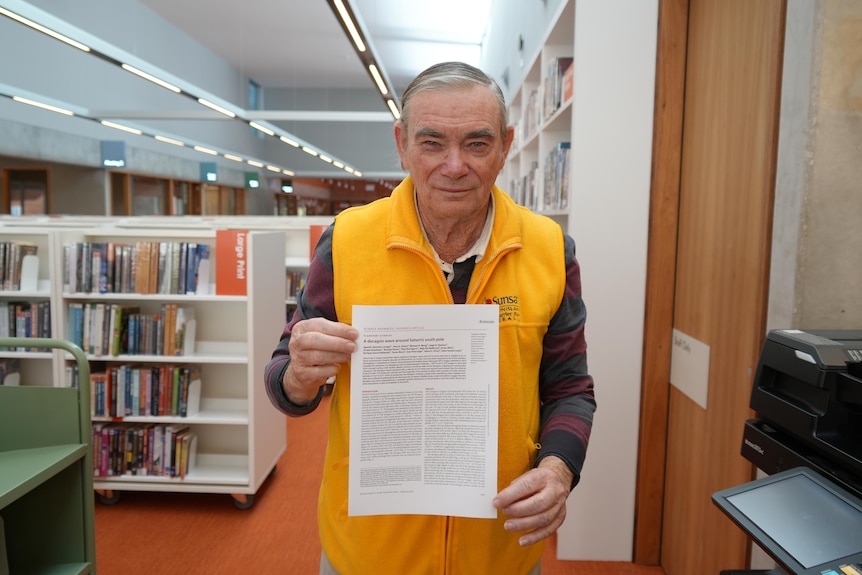 Man holds research paper in library next to printer and in front of shelf of books 