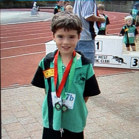 A boy smiles at a running track, wearing two medals and school uniform.