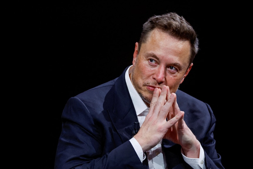 Elon Musk leans forwards with his hands pressed together, resting on his face