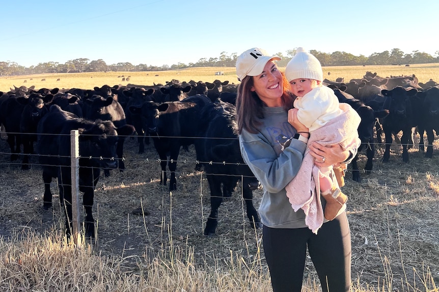 A woman stands in front of a herd of angus cattle holding a baby daughter