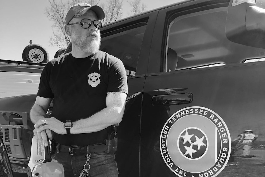 A man in jeans, a black t-shirt, cap and sunglasses standing next to a black SUV with a circle logo on the door.