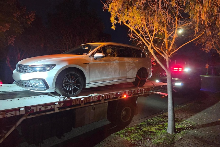 A white sedan on the tray of a tow truck parked in front of a police car