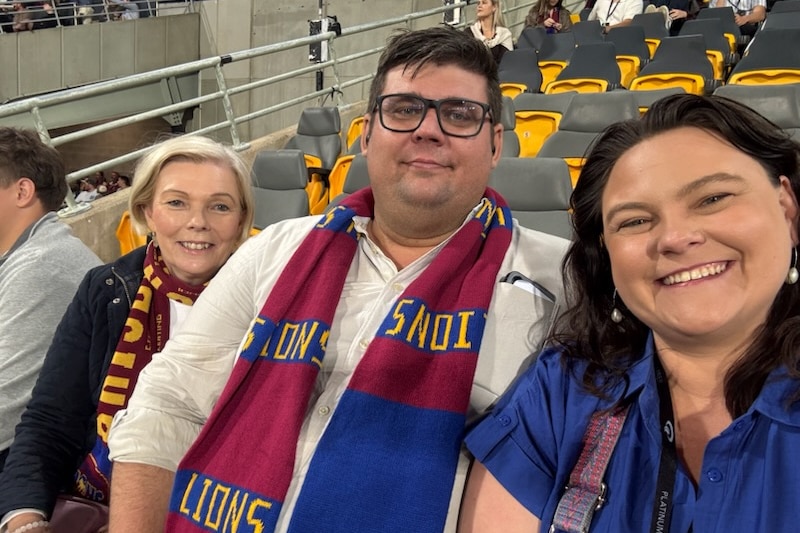 A middle-aged blonde woman with a dark-haired man and a dark-haired woman in the stands at a stadium.
