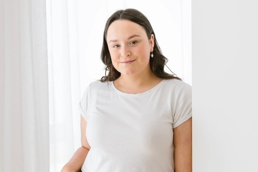 A young woman with brown hair wearing a white top and blue jeans leans against a wall, smiling
