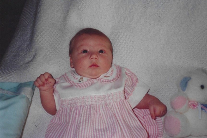 A baby in a pink and white dress laying on a bed with a stuffed bear next to them.