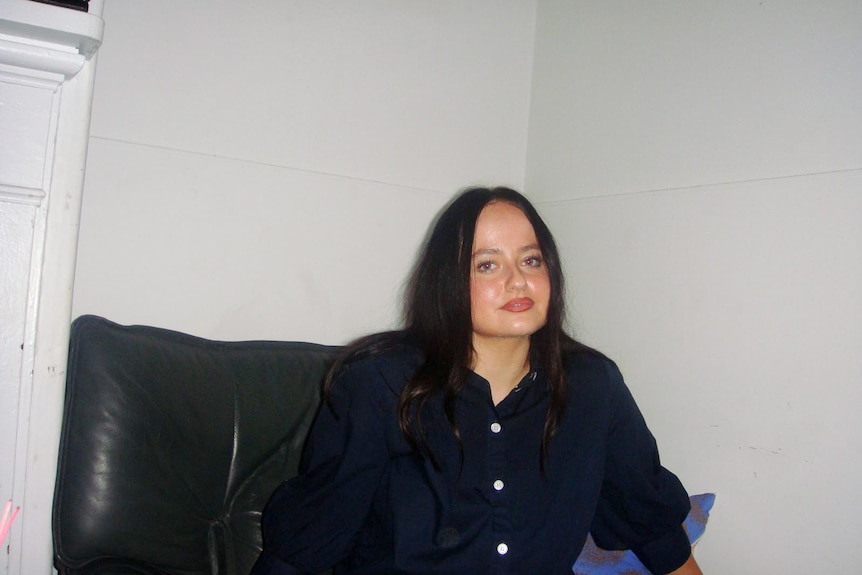 A young woman with brown hair sits on a couch, smiling