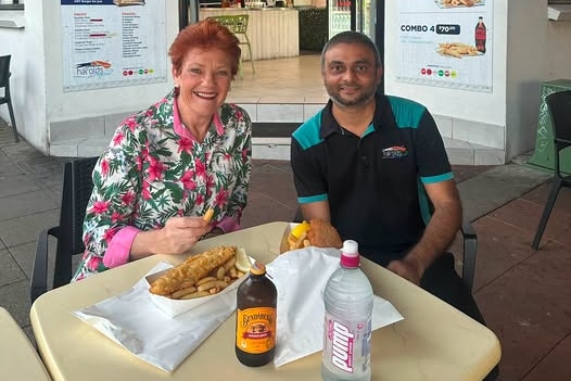 A woman in a floral patterned collared shirt eating fish and chips sitting with a man in a black and cyan collared t shirt.