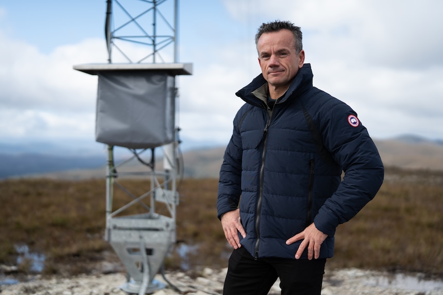 Stephen Casey wearing navy puffer jacket, stands with hands on hips on top of a hill