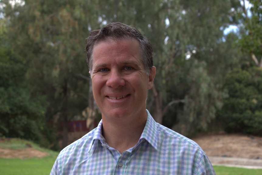 A smiling man (Justin Clancy) wearing an open-collared business shirt stands in front of some trees, looking at the camera.