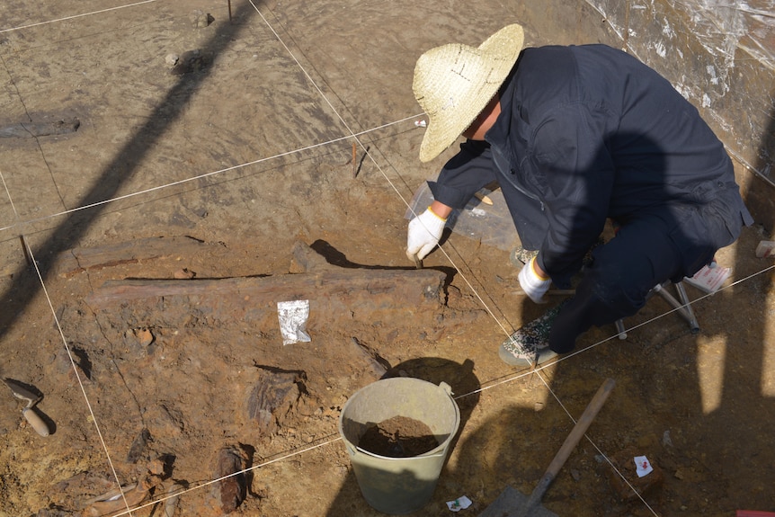 Person wearing hat digging things out of dirt floor in cave.