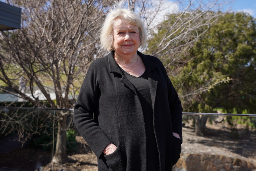 A woman in black clothes stands in front of trees.