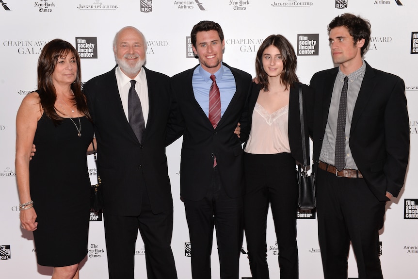 A couple stand with their three adult children. All are wearing smart trouser suits. The mother is wearing a black dress.