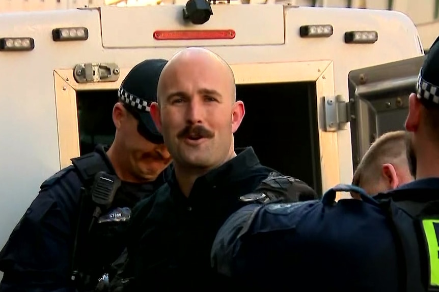 A bald man with a brown moustache looks at the camera while being handcuffed by police.