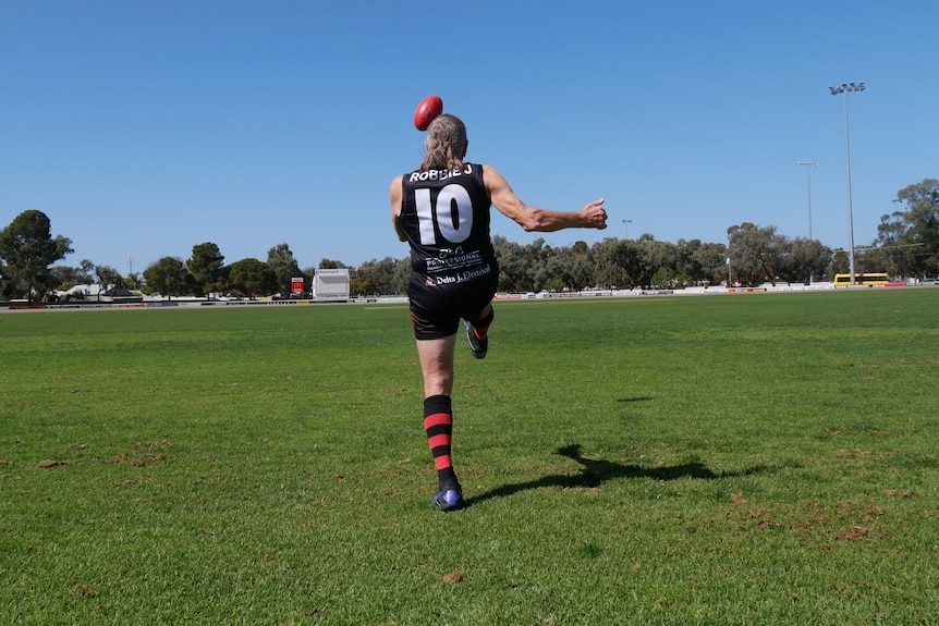 An elderly man seen from the back in a football guernsey with number 10 on it kicks a red Australian Rules football.