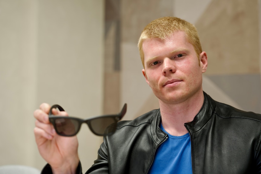 A man wearing a black leather jacket over a blue t-shirt holds a pair of black sunglasses he has short cropped light hair