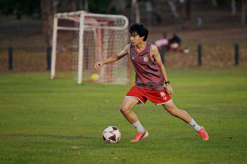 Mark O'Connor, wearing a red and grey patterned training jersey, kicks a ball across a grass field with a set of goals behind.