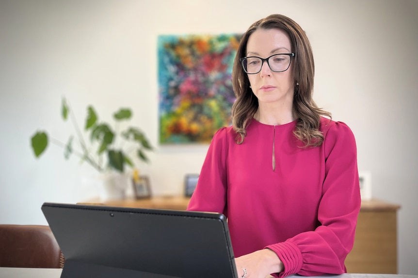 A white woman with long brown hair and glasses working on a laptop