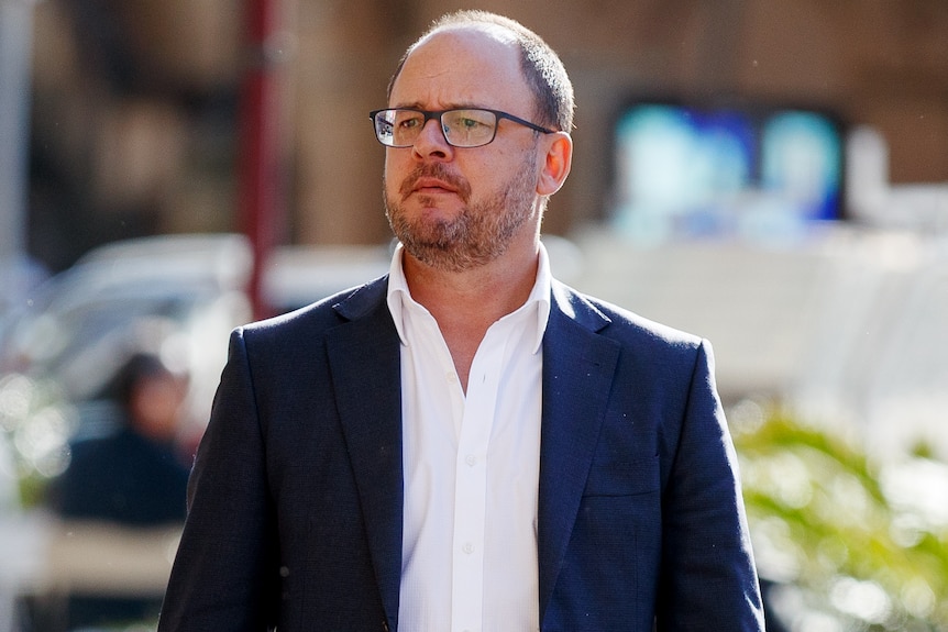 A middle-aged man in glasses and a dark suit stands in the sun.